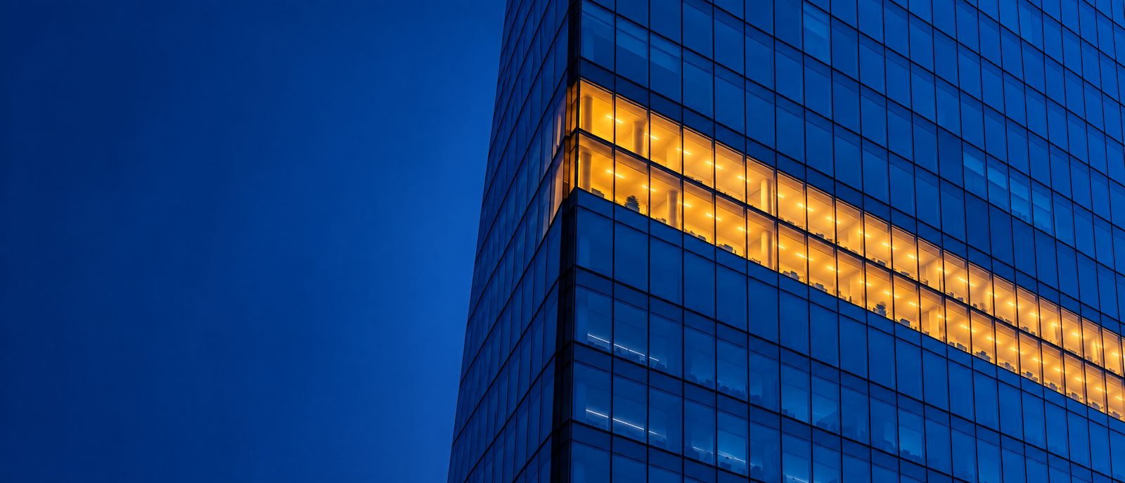 Fachada de vidrio de una torre corporativa en la hora azul, con una franja de pisos iluminada en ámbar