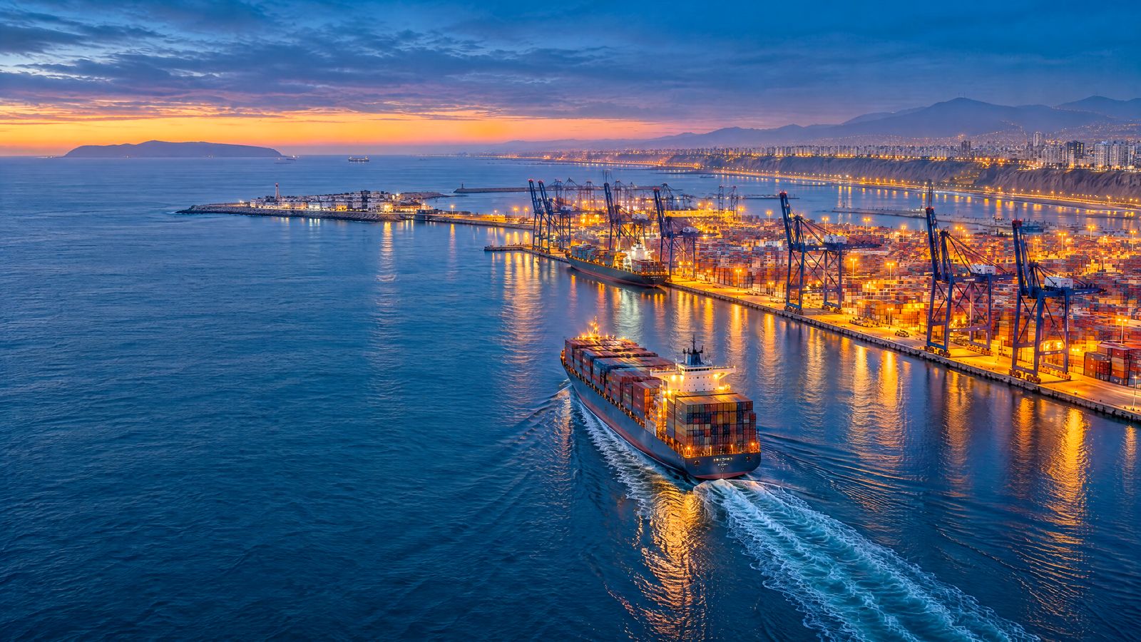 Vista aérea del terminal de contenedores del puerto del Callao en la hora azul, con un buque saliendo hacia el Pacífico, la isla San Lorenzo y las luces de Lima al fondo